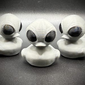 May include: Three white alien-themed rubber ducks with large black oval eyes. The ducks are arranged in a row, each with a rounded body and a duck-like bill. The ducks are a novelty item.