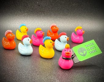 May include: A collection of colorful duck figurines with various crest colors, including pink, yellow, and orange. One duck has a black collar and a tag that reads "TRUTH IS OUT THERE" with an alien graphic. The ducks are on a dark surface.