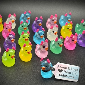 May include: A collection of colorful duck figurines in various shades, including pink, green, yellow, blue, and purple. Each duck has a small, pink horn-like detail on its head and an orange beak. A tag reads "Peace & Love from Oklahoma."
