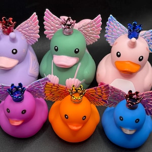 May include: A collection of six colorful rubber ducks, each adorned with iridescent wings and a miniature crown. The ducks come in various colors, including purple, green, pink, magenta, orange, and blue, creating a whimsical and eye-catching display.