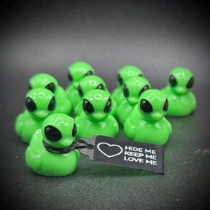 May include: A collection of green alien-themed rubber ducks with black eyes. One duck holds a black tag with a white heart and the text "HIDE ME KEEP ME LOVE ME". The ducks are arranged on a dark surface.
