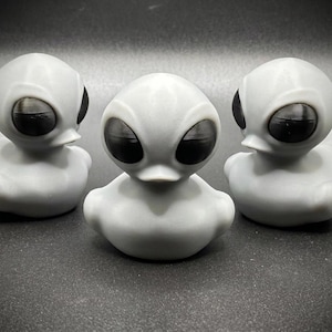 May include: Three gray alien-themed rubber ducks with large black eyes. The ducks are arranged in a row, each with a rounded body and a duck-like bill. The background is a dark gray.
