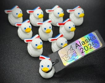 May include: A collection of white duck figurines with blue eyes and yellow beaks. Each duck has a red, white, and blue accent on its head. A tag with the text "Alaska 2025" is attached to one of the ducks.