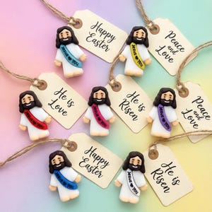 Può includere: Una collezione di piccole statuette di Gesù con capelli e barba castani, che indossano vesti bianche e fasce colorate. Ogni statuetta è abbinata a un'etichetta con scritto "Buona Pasqua", "Lui è Amore", "È risorto" o "Pace e Amore".