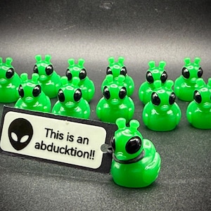 May include: A group of green alien-themed rubber ducks with black eyes and antennae. One duck holds a tag that reads "This is an abducktion!!" with an alien head graphic. The ducks are arranged on a dark surface.