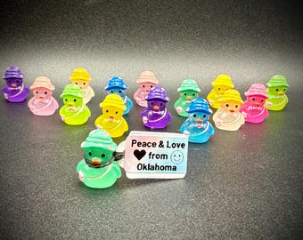 May include: A collection of colorful duck figurines, each wearing a hat and scarf. The ducks are in various colors, including purple, pink, green, yellow, and blue. A tag reads "Peace & Love from Oklahoma."