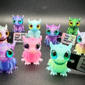 May include: A selection of small dragon figurines in various colours, including blue, purple, pink, and green. Each dragon has large eyes and is displayed on a dark surface. The tags attached to the dragons read "Tiny but Mighty."