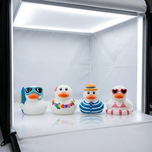 Puede incluir: Cuatro patitos de goma blancos con diferentes accesorios. Uno lleva gafas de sol azules, otro un collar de flores, un tercero una camisa a rayas y un sombrero, y el último gafas de sol en forma de corazón y un flotador rosa.