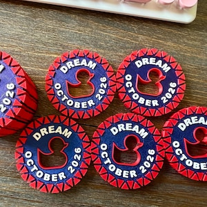 May include: Six circular tokens featuring a dark blue center with a red rubber duck silhouette, the text DREAM OCTOBER 2026 in white, and a red serrated border. One token is stacked on top of others, while five are arranged flat in a semi-circle.