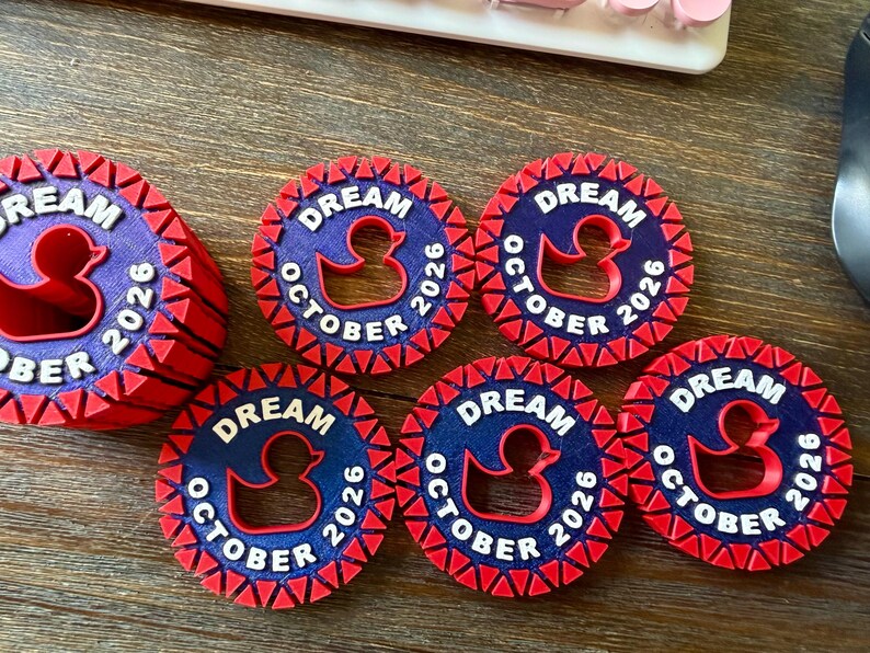 May include: Six circular tokens featuring a dark blue center with a red rubber duck silhouette, the text DREAM OCTOBER 2026 in white, and a red serrated border. One token is stacked on top of others, while five are arranged flat in a semi-circle.
