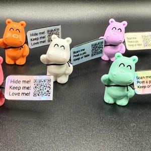 May include: A collection of colorful hippo figurines in orange, pink, white, purple, and green. Each hippo holds a small sign with a QR code and text that reads "Hide me! Keep me! Love me!" or "Scan me, Post a pic, Keep or hide."