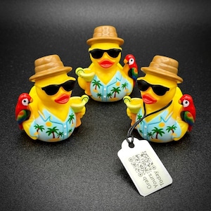 Peut inclure: Trois canards en caoutchouc jaunes portant des lunettes de soleil, des chapeaux bruns et des chemises hawaïennes, chacun tenant un cocktail. Un perroquet coloré est assis sur l'épaule de chaque canard. Une étiquette blanche avec le texte "Grab Yours Today" est suspendue à l'un des canards.