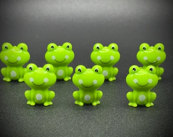 Puede incluir: Siete pequeñas figuras de ranas verdes sonrientes con manchas blancas y ojos negros están dispuestas sobre una superficie oscura. Las ranas tienen un diseño de dibujos animados, con una expresión alegre.
