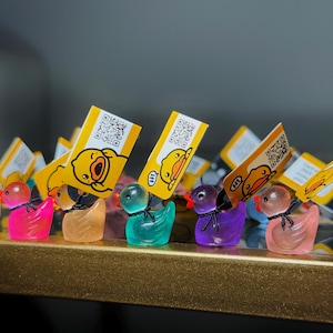 May include: A collection of colorful, translucent duck figurines. Each duck has a small black bow and a QR code tag with a cartoon duck illustration. The ducks are in shades of pink, orange, teal, purple, and peach.