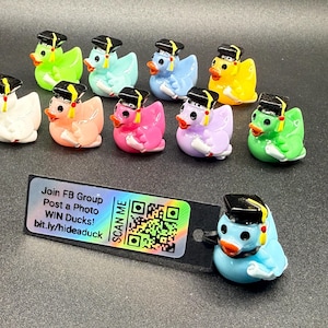 Può includere: Una collezione di figurine di paperelle di gomma colorate, ognuna con un cappello di laurea nero con un nappa gialla. Le paperelle sono di vari colori, tra cui bianco, rosa, blu, verde e giallo. È presente anche un'etichetta con testo e un codice QR.
