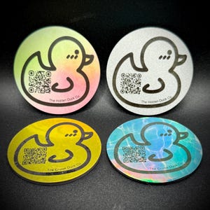 Peut inclure: Quatre autocollants circulaires avec un motif de canard de dessin animé et un code QR. Les autocollants présentent différents schémas de couleurs : un dégradé de vert, jaune et rose ; argent ; jaune ; et bleu avec un effet d'eau. Le texte "The Hidden Duck Co" et "The Cruise Duck Co" est imprimé.