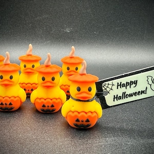 May include: A group of yellow and orange Halloween duck figurines, each with a pumpkin-shaped body and a matching hat. A small sign with the words "Happy Halloween!" and a bat illustration is also present.