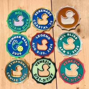 May include: Nine circular, scalloped-edge plastic tags featuring a central rubber duck cutout. Text includes NCL CRUISIN DUCK, SERENADE COASTAL '26, SUMMER BASH 2026, LEGEND AUG 9-20, 2026, NCL BLISS APR 5, 2026, SYMPHONY CRUISE 2026, and VENEZIA 2/21-3/7/27.