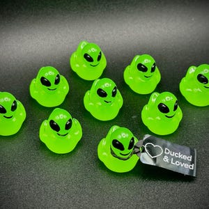 Può includere: Una collezione di dieci paperelle di gomma a forma di alieno verde neon con occhi e sorrisi neri. Una paperella ha un'etichetta con la scritta "Ducked & Loved". Le paperelle sono disposte su una superficie scura.
