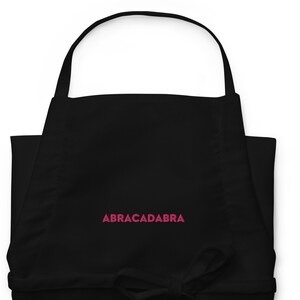 Puede incluir: Delantal negro con tirantes ajustables y un texto bordado en rosa "ABRACADABRA".