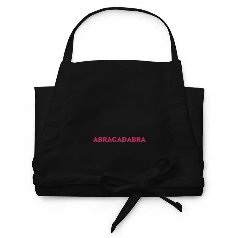 Puede incluir: Delantal negro con tirantes ajustables y un texto bordado en rosa "ABRACADABRA".