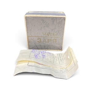 May include: A rectangular gray box with a textured surface featuring the Cyrillic text "ЧАСЫ" and "ЗАРЯ" in gold lettering. Accompanying the box is a folded, translucent paper document with printed Cyrillic text and a blue ink stamp dated 1976.