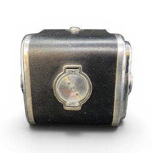 May include: A vintage black leather-covered camera body with silver metal trim. The front features a circular light meter dial with the Cyrillic text GOST printed in the center, surrounded by numerical markings and a small red icon at the bottom.