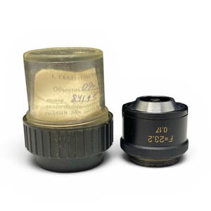 May include: Two vintage camera lenses. One lens is in a clear plastic case with a paper label. The other lens is black with gold lettering, including "F=23.2 0.17".