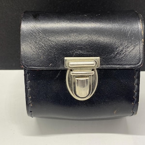 May include: A small, black leather case with a silver-colored clasp. The case is rectangular with a hinged lid and visible stitching along the edges. The clasp is centered on the front, providing a secure closure.