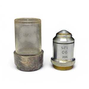 May include: A microscope objective lens with a clear plastic case. The lens is silver with gold accents and has the numbers "90" and "1.25" printed on it. The case is clear with a marbled base.
