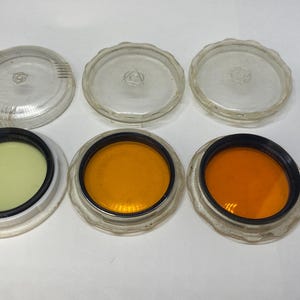 May include: A collection of six vintage camera lens filters. Three filters are clear with a ridged edge, and three are colored: yellow-green, orange, and red-orange. The filters are in clear plastic cases.