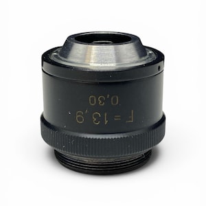 May include: A black and silver microscope objective lens. The lens has a knurled ring at the base and a silver top. The text "F=13.9, 0.30" is etched on the side of the lens.