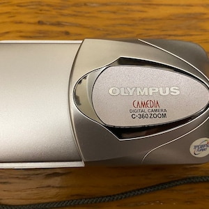 May include: A silver Olympus Camedia digital camera with a zoom lens. The camera has a black strap and a small blue and white sticker that says "Zoom Lens".