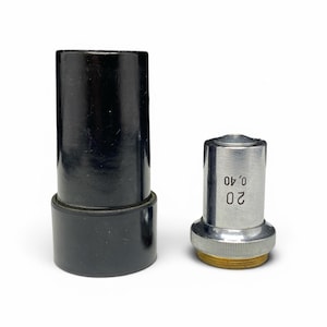 May include: Two microscope objective lenses. One is a black cylindrical tube with a wider base. The other is a silver-colored lens with the numbers "20 0,40" printed on the side and a gold-colored base.