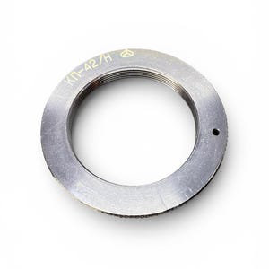 Op de afbeelding: Een cirkelvormige metalen camera-lensadapterring met een donkergrijze afwerking. Het bovenoppervlak bevat gegraveerde gele tekst KP-42/H en een klein driehoekig logo. De binnenrand is voorzien van schroefdraad en een klein gaatje is zichtbaar.