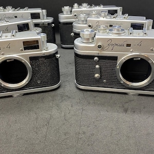 Op de afbeelding: Verschillende vintage Zorki-4 camera's zijn gerangschikt op een donkere ondergrond. De camera's hebben een zilveren bovenkant en een zwarte, getextureerde behuizing. De lens is een opvallende cirkelvormige opening. De naam "Зоркий -4" is gedrukt.