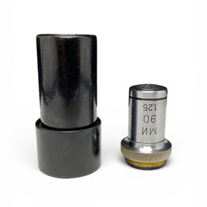 May include: A black cylindrical container next to a silver microscope objective. The objective has the numbers "90" and "1.25" printed on it. The objective has a gold-colored ring at the base.