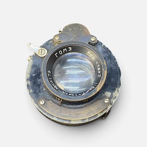 May include: A vintage circular camera lens assembly with a weathered black and brass finish. The lens features Cyrillic text including GOMZ and Industar, with technical markings for aperture and shutter speed settings visible on the metal housing.