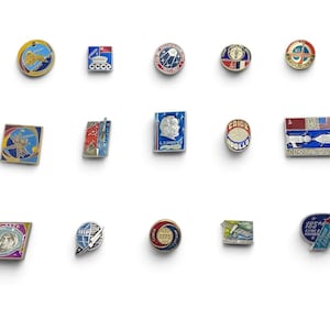 May include: A collection of fourteen metal enamel pins featuring space exploration themes, including Soviet-era designs, rocket imagery, astronaut portraits, and mission emblems in various shapes like circles, squares, and rectangles with blue, red, and gold.