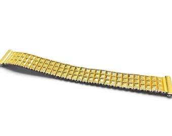 May include: A gold-toned metal watch band featuring a flexible, expandable link design. The surface of the band is composed of a repeating pattern of small, raised square segments, creating a textured appearance across the entire length of the strap.