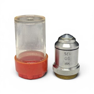 May include: A microscope objective lens and its protective case. The lens is silver with black text that reads "90 1.25 MM". The case is clear plastic with a red base.