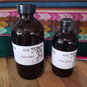 May include: Two amber glass bottles with black lids and white labels. The labels have a black and white floral design and the text "Fire Cider". The larger bottle is labeled "8 fl oz" and the smaller bottle is labeled "4 fl oz".
