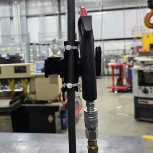 May include: A black air gun with a red trigger is attached to a black hose. The hose is secured to a dark gray pole with black brackets and white zip ties. The background shows industrial equipment.