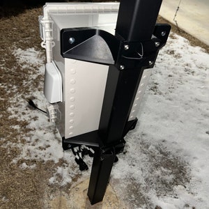 May include: A white weatherproof box mounted to a black metal pole with black plastic brackets. The box has a white plastic cover and a black power cord.