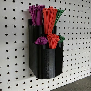 May include: A black 3D-printed organizer mounted on a pegboard, holding bundles of purple, orange, and green cable ties. The organizer has multiple compartments for easy access and storage of various sizes of zip ties.