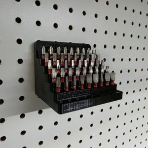 May include: A black tiered organizer filled with various metal and red-tipped drill bits. The organizer is mounted on a white pegboard with evenly spaced holes. The bits are arranged in rows.