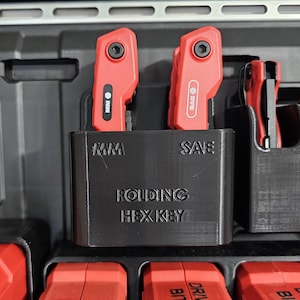 May include: A close-up of a black organizer with red tools. The organizer has the text "MM", "SAE", and "FOLDING HEX KEY". Two red folding hex keys are in the center, with a red tool on the right. The tools are in a black case.