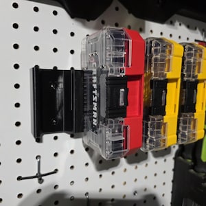 May include: Three clear plastic tool organizers with yellow and red lids. The organizers are mounted on a white pegboard wall. The organizers are labeled "CRAFTSMAN".