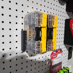 May include: Two yellow and clear plastic tool boxes with the word "DEWALT" printed on the side, mounted on a white pegboard wall.