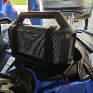 May include: A black portable Bluetooth speaker with a gray speaker grill and a black strap. The speaker is mounted on a blue vehicle.